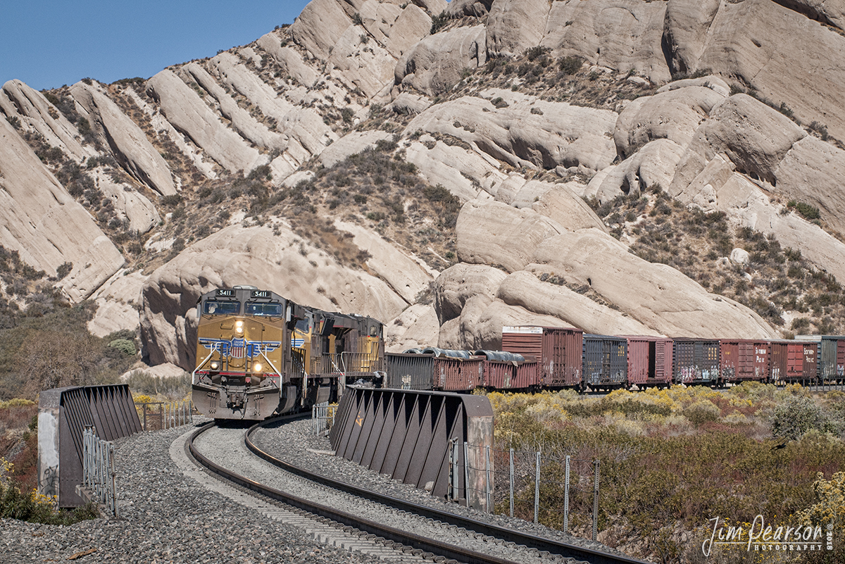 October 20, 2008 - Union Pacific 5411 leads a mixed freight westbound past Mormon Rocks in the southern California's famous Cajon Pass. 

According to Wikipedia: Cajon Pass (/k?'ho?n/; elevation 3,777 ft (1,151 m) is a mountain pass between the San Bernardino Mountains and the San Gabriel Mountains in Southern California in the United States. It was created by the movements of the San Andreas Fault. Located in the Mojave Desert, the pass is an important link from the Greater San Bernardino Area to the Victor Valley, and northeast to Las Vegas.

Cajon Pass is at the head of Horsethief Canyon, traversed by California State Route 138 (SR 138) and railroad tracks owned by BNSF Railway and Union Pacific Railroad. Railroad improvements in 1972 reduced its maximum elevation from about 3,829 to 3,777 feet (1,167 to 1,151 m) while also reducing the curvature. Interstate 15 does not traverse Cajon Pass, but rather the nearby Cajon Summit,  elevation 4,260 feet (1,300 m). However, the entire area including Cajon Pass and Cajon Summit is often collectively called Cajon Pass. Sometimes the entire area is called Cajon Pass, but a distinction is made between Cajon Pass and Cajon Summit in detail.

Mormon Rocks
In 1851, a group of Mormon settlers led by Amasa M. Lyman and Charles C. Rich traveled through the Cajon Pass in covered wagons on their way from Salt Lake City to southern California. This prominent rock formation in the pass, where the Mormon trail and the railway merge (near Sullivan's Curve), is known as Mormon Rocks.