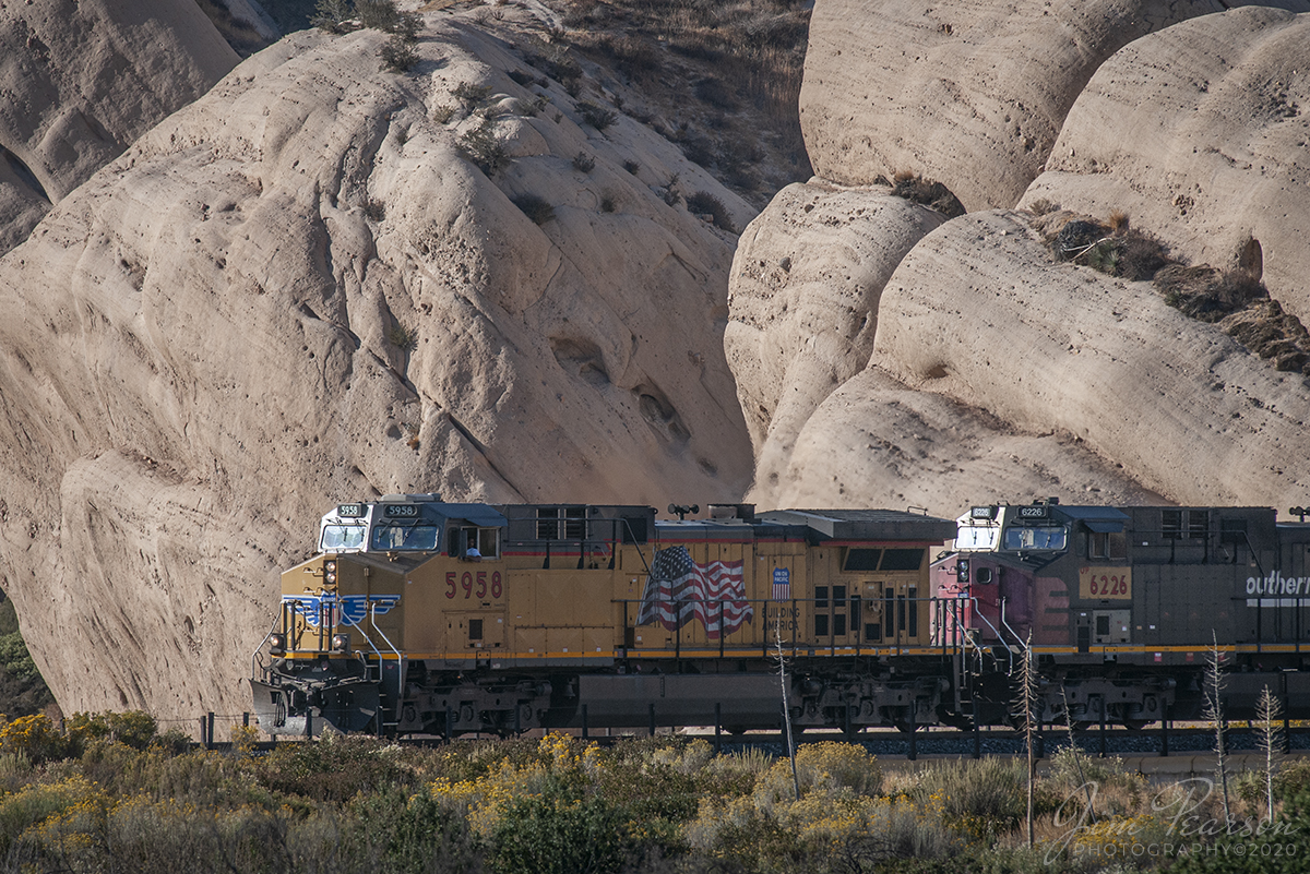 October 20, 2008 - The late evening light rakes across the base of Mormon Rocks as UP 5958 and 6266 (ex-Southern Pacific Unit) lead a west bound freight down the Cajon Pass in southern California. 

Local legend has it that the rocks were named for the Mormon pioneers who camped here after their descent from the pass ridgeline. Another legend calls the rocks the 'Chanting Rocks,' as when the wind would blow across the portholes in the rocks it was said the sound made was similar to a low chanting or singing.