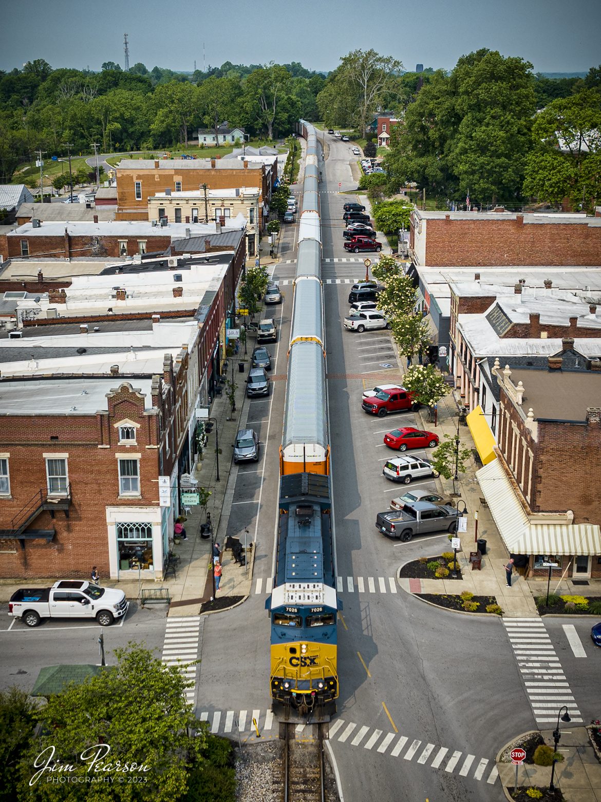CSXT 7026 leads a loaded autorack train as they street run through downtown LaGrange, Kentucky, headed north on the LCL Subdivision on May 17th, 2023.

According to the Train Aficionado website: CSX runs through La Grange on what is known as the LCL Subdivision. This 101-mile line connects Covington, Kentucky, to Louisville, Kentucky. The railroad tracks run right through the heart of the town literally. The railroad tracks are embedded on the eastbound side of the roadway for the whole stretch of downtown. The trains pass the city business district which includes quaint shops and restaurants. When looking up railfanning in La Grange most websites state anywhere from 14 to 20 trains travel through here daily.

According to Hidden History of La Grange, Ky by author and local historian Nancy Stearns, LaGrange is known for its trains. It became an important junction for the railroad, with lines going to Frankfort and Cincinnati, and this made the town an important stop for travelers and businesses. The interurban electric rail car made round trips from LaGrange to Louisville daily until automobiles surpassed the interurban in popularity.

Tech Info: DJI Mavic 3 Classic Drone, RAW, 24mm, f/2.8, 1/1250, ISO 100.

#trainphotography #railroadphotography #trains #railways #dronephotography #trainphotographer #railroadphotographer #jimpearsonphotography #CSXtrains #mavic3classic #drones #trainsfromtheair #trainsfromadrone #emptycoaltrain #autoracktrain #kentuckytrains #LaGrangeKy #streetrunningtrains