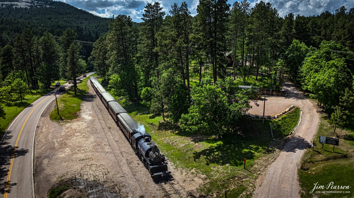A local artist gives a friendly wave from her driveway as the Black Hills Central Railway locomotive 108 heads to Keystone, South Dakota, on June 26th, 2025. A big shout out to Cory Jakeway for all the help on finding my way around on the railroad and railfanning with me!

According to their website: Locomotive is a 2-6-6-2T articulated tank engine that was built by the Baldwin Locomotives Works in 1926 for the Potlatch Lumber Company. It later made its way to Weyerhaeuser Timber Company and eventually to the Northwest Railway Museum in Snoqualmie, Washington.

The acquisition and subsequent restoration of locomotive 108 completed a more than 20-year goal of increasing passenger capacity which began with the restoration of 110 and the restoration of multiple passenger cars. Both large Mallet locomotives (pronounced “Malley”) can pull a full train of seven authentically restored passenger cars, up from the four cars utilized prior to their addition to the roster.

Tech Info: DJI Mavic 3 Classic Drone, RAW, 24mm, f/2.8, 1/4000, ISO 130.

#railroad #railroads #train, #trains #railway #railway #steamtrains #railtransport #railroadengines #picturesoftrains #picturesofrailways #besttrainphotograph #bestphoto #photographyoftrains #bestsoldpicture #JimPearsonPhotography #trainsfromtheair #trainsfromadrone #1880train #steamtrain #blackhills