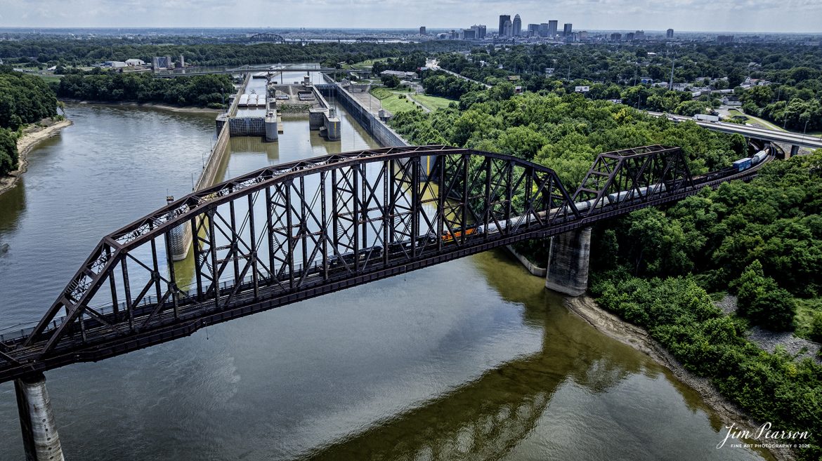Norfolk Southern 7762 leads NS 168 westbound over the Kentucky & Indiana Bridge (K&I bridge) out of Louisville, Kentucky, over the Ohio River into New Albany, Indiana on the NS Louisville District, on July 28th, 2025. From what I can tell the third unit is an ex-BNSF locomotive. In the background is the lock for barge traffic and the city of Louisville, Ky.

According to Wikipedia: The Kentucky & Indiana Bridge is one of the first multi modal bridges to cross the Ohio River. It is for both railway and common roadway purposes together. By federal, state, and local law railway and streetcar, wagon-way, and pedestrian modes of travel were intended by the City of New Albany, City of Louisville, State of Kentucky, State of Indiana, the United States Congress, and the bridge owners.

The K & I Bridge connects Louisville, Kentucky to New Albany, Indiana. Constructed from 1881 to 1885 by the Kentucky and Indiana Bridge Company, the original K&I Bridge opened in 1886. It included a single standard gauge track and two wagon ways, allowing wagons and other animal powered vehicles to cross the Ohio River by a method other than ferry for the first time. At the time motorized vehicles were virtually nonexistent.

The K&I Bridge company also owned a ferry boat operation during both the 1st and 2nd bridge; eventually that operation was sold as the bridge’s success largely outmoded boat usage.

In 1910 the bridge company was renamed the Kentucky & Indiana Terminal Railroad Co. From 1910 to 1912, a new, heavier bridge was built on new piers just upstream from the original one, after which the old bridge was demolished. The new bridge was double tracked to handle increasingly heavier train and now automobile traffic, eventually receiving the U.S. 31W designation.

The bridge also featured a rotating swing span opening for the passage of ships in high water. The bridge was only opened four times, twice for testing in 1913 and 1915, then in 1916 for the passage of the steamer “Tarascon” and in 1920 for passage of the Australian convict ship “Success”. In 1948 it refused opening of the span for passage of the steamer “Gordon C. Greene” citing inconvenience and costs of cutting power and communication lines, an action for which K&I and LG&E both paid damages to that ship’s company. In 1955 the K&I sought and received permission to permanently tie down the swing span from the Corps of Engineers. In 1952, the creosoted wood block roadways of the second bridge were eliminated and replaced by a steel gridwork roadway.

On February 1, 1979, an overweight dump truck caused a small segment of the steel grate roadway on the bridge to sag about 1 foot (0.30 m). A quick survey promised to reopen the roadway, but automotive traffic was banned thereafter by the railroad.

Tech Info: DJI Mavic 3 Classic Drone, RAW, 22mm, f/2.8, 1/1600, ISO 130.

#railroad #railroads #train, #trains #railway #railway #steamtrains #railtransport #railroadengines #picturesoftrains #picturesofrailways #besttrainphotograph #bestphoto #photographyoftrains #bestsoldpicture #JimPearsonPhotography #trainsfromtheair #trainsfromadrone