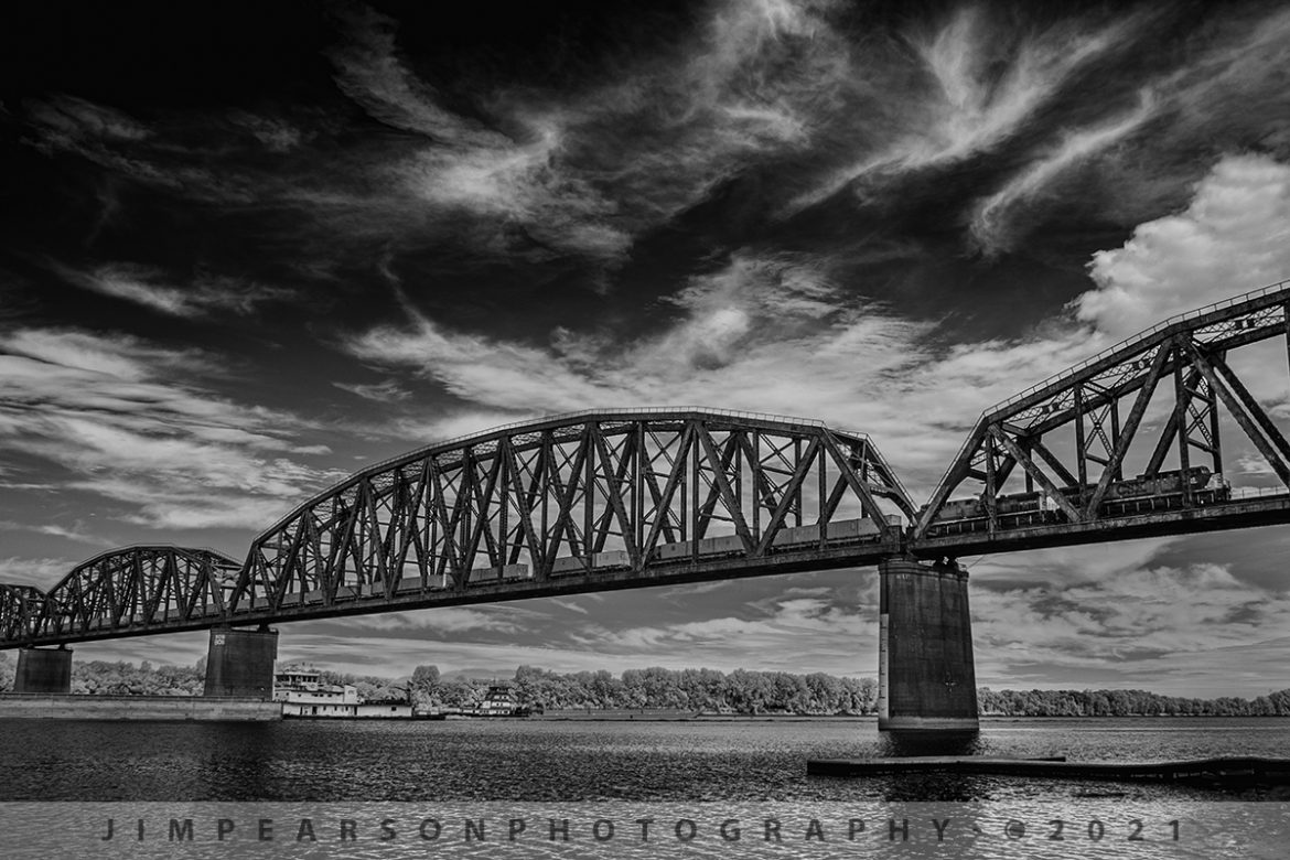 Trains and barges on and over the Ohio River at Henderson, KY

In this Infrared shot from August 13th, 2021, my plan was to catch two barges passing each other under the bridge on the Ohio River at Henderson, Kentucky while I was waiting for military train CSX W816 to make its way south on the Henderson Subdivision. 

I never expected the train to head over the river during this moment, as I hadnt heard it call the ballast deck (last place they call on the scanner before hitting the bridge) before I got out of the car and decided to grab the barge shot in infrared while I was waiting. If I had known, it was this close I would have put my drone up for this shot! Not that I'm complaining at all though!

The containers behind the power contain the munitions for the last half of the train, which was armor, consisting of Bradley Fighting Vehicles and M1 Tanks.

Tech Info: Fuji XT-1, RAW, Converted to 720nm B&W IR, Fuji 18-55 @18mm, f/3.6, 1/500, ISO 200.