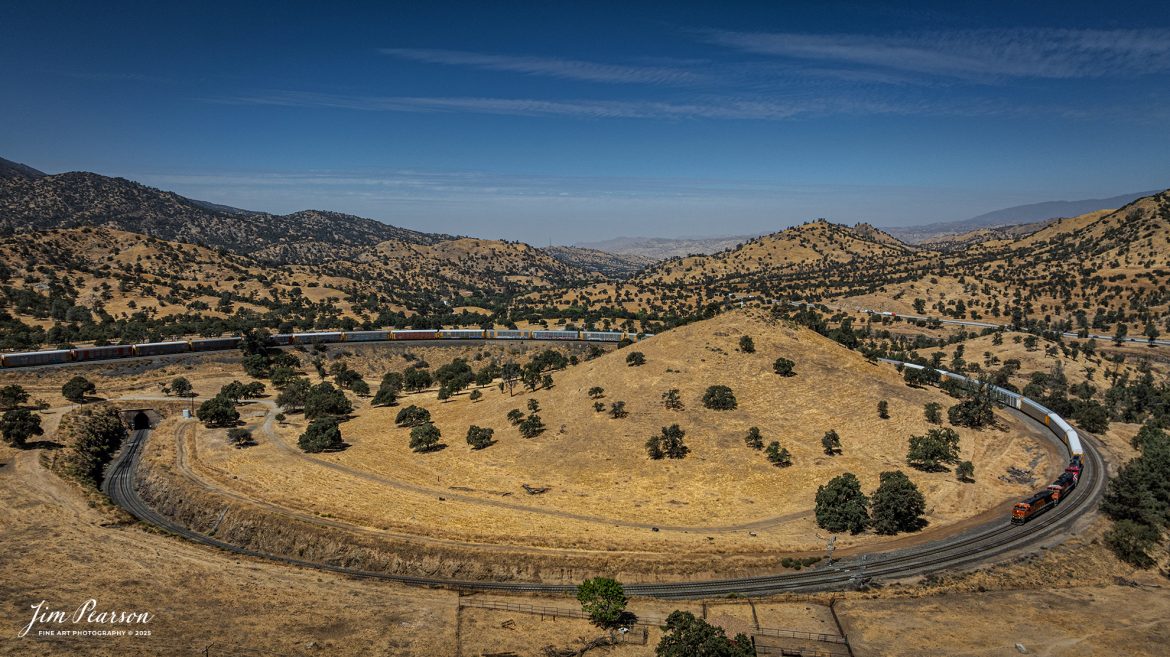 September 18th, 2024, BNSF 7115, 9128 and Ferromex 4050 lead an northbound train through Tehachapi Loop on the Union Pacific Mojave Subdivision at Keene, California.

According to Wikipedia: The Tehachapi Loop is a 3,779-foot-long (0.72 mi; 1.15 km) spiral, or helix, on the Union Pacific Railroad Mojave Subdivision through Tehachapi Pass, of the Tehachapi Mountains in Kern County, south-central California. The line connects Bakersfield and the San Joaquin Valley to Mojave in the Mojave Desert.

Rising at a steady two-percent grade, the track gains 77 feet (23 m) in elevation and makes a 1,210-foot-diameter (370 m) circle. Any train that is more than 3,800 feet (1,200 m) long—about 56 boxcars—passes over itself going around the loop. At the bottom of the loop, the track passes through Tunnel 9, the ninth tunnel built as the railroad was extended from Bakersfield.

The line averages about 36 freight trains each day. Passenger trains such as Amtrak's San Joaquin are banned from the loop, although the Coast Starlight can use it as a detour. Its frequent trains and scenic setting make the Tehachapi Loop popular with railfans. In 1998, it was named a National Historic Civil Engineering Landmark. It is also designated as California Historical Landmark #508.

One of the engineering feats of its day, the Loop was built by Southern Pacific Railroad to ease the grade over Tehachapi Pass. Construction began in 1874, and the line opened in 1876.

Tech Info: DJI Mavic 3 Classic Drone, RAW, 24mm, f/2.8, 1/2500, ISO 100.

#railroad #railroads #train, #trains #railway #railway #steamtrains #railtransport #railroadengines #picturesoftrains #picturesofrailways #besttrainphotograph #bestphoto #photographyoftrains #bestsoldpicture #JimPearsonPhotography #trainsfromtheair #trainsfromadrone #TehachapiLoop