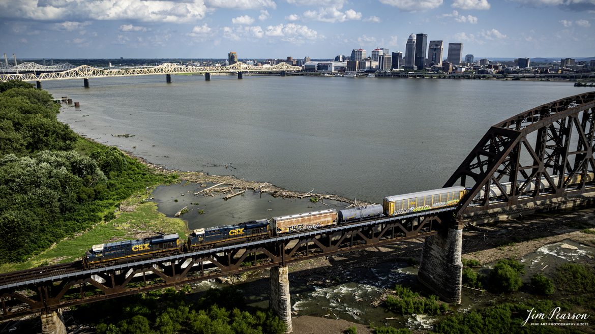 CSX M688 heads north across the Ohio River on the 14th Street Bridge (Louisville and Indiana (L&I) Bridge) at Louisville, Ky on July 28th, 2025. This bridge carries traffic for the Louisville and Indiana, along with the CSX Railroad.In the background is downtown Louisville, Ky.

According to Wikipedia: The Fourteenth Street Bridge, also known as the Ohio Falls Bridge, Pennsylvania Railroad Bridge, Conrail Railroad Bridge or Louisville and Indiana (L&I) Bridge, is a truss drawbridge that spans the Ohio River, between Louisville, Kentucky and Clarksville, Indiana.

Built by the Louisville Bridge Company and completed in 1870, the bridge was operated for many years by the Pennsylvania Railroad, giving the company its only access to Kentucky. Ownership of the railroad and the bridge passed on to Penn Central and later Conrail, which then sold the line from Louisville to Indianapolis, Indiana to the Louisville and Indiana Railroad, the current bridge owner.

The draw portion of the bridge is a vertical-lift span, built in about 1918 in place of a swing span. The towers and machinery of the lift span were designed by Waddell and Son, Inc., and there is a plaque on the SW tower reading, "Waddell Vertical Lift Bridge, Waddell and Son, 1917". The draw span is across the upstream end of the Louisville and Portland Canal, which includes the McAlpine Locks and Dam. Ohio River traffic passes through this canal to navigate past the Falls of the Ohio. 

Tech Info: DJI Mavic 3 Classic Drone, RAW, 22mm, f/2.8, 1/2000, ISO 110.

#railroad #railroads #train, #trains #railway #railway #steamtrains #railtransport #railroadengines #picturesoftrains #picturesofrailways #besttrainphotograph #bestphoto #photographyoftrains #bestsoldpicture #JimPearsonPhotography #trainsfromtheair #trainsfromadrone